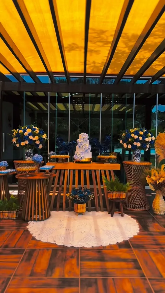 Celebração sob cobertura iluminada, com bolo e arranjos de flores azuis e amarelas